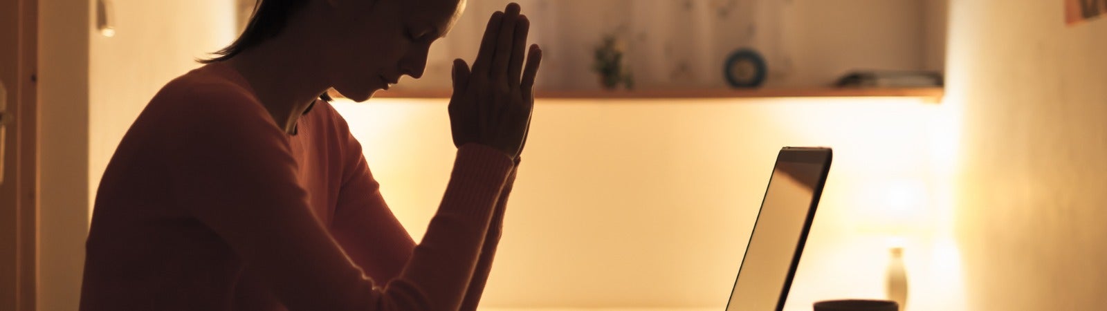 A woman sits and prays in front of her laptop.