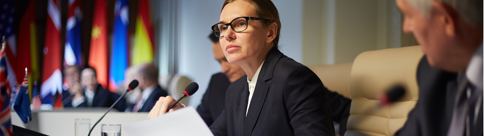 woman seated at conference table with microphone and flags