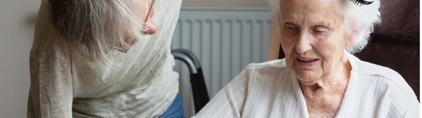 A caregiver (left) bends down and brushes the hair of an older woman (right) who is seated. They both have gray hair and the caregiver is wearing glasses.