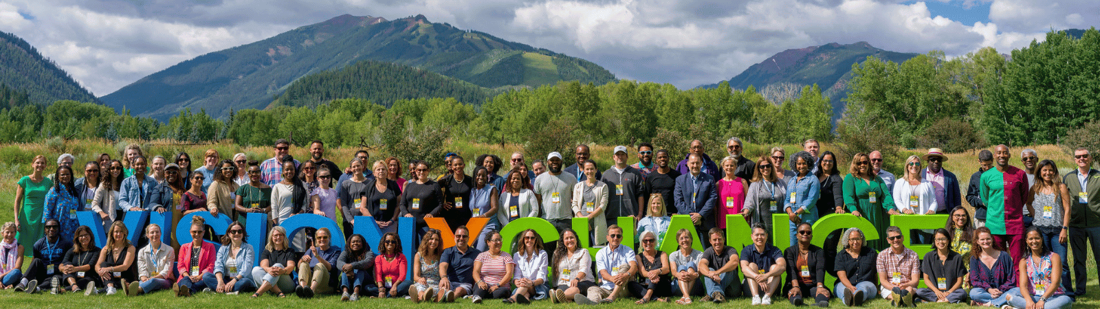 A large group of event participants stand and sit together, smiling. They are on a grassy area and behind them are the rocky mountains and a cloudy sky.