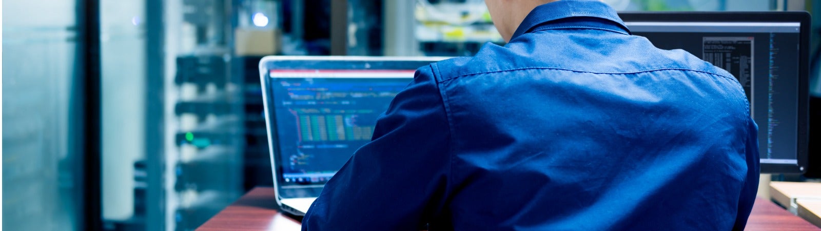 A man sits at a computer in a server room looking at lines of code.