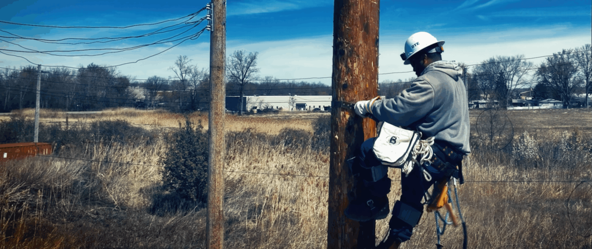 A utility worker in a safety harness climbs a wooden power pole in a rural area, wearing a white hard hat and gray hoodie, with electrical wires visible above and dry grassy fields in the background.