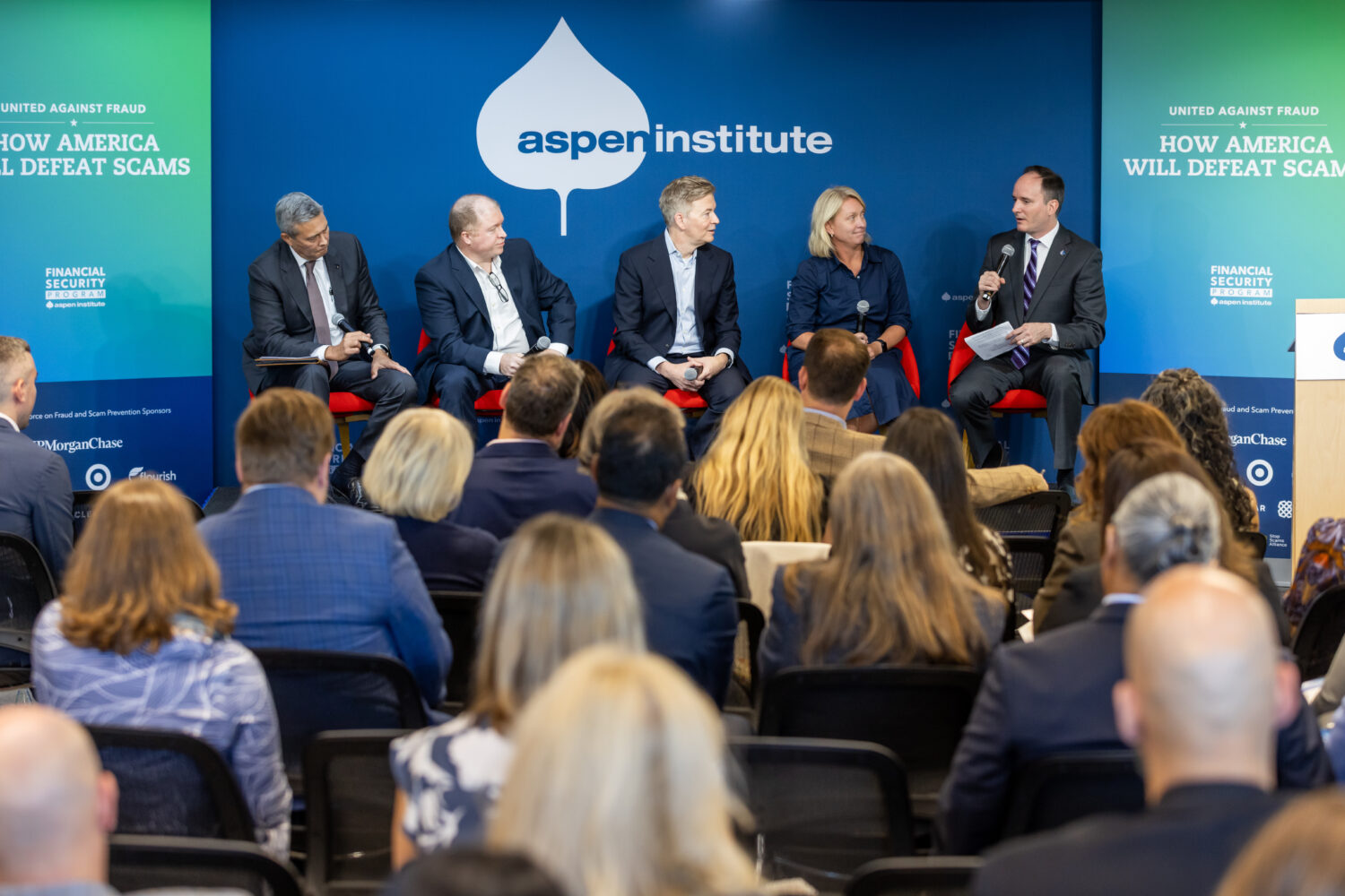 Five panelists sit before a large crowd. In the background a banner reads "How America Will Defeat Scams" with the Aspen Institute logo.