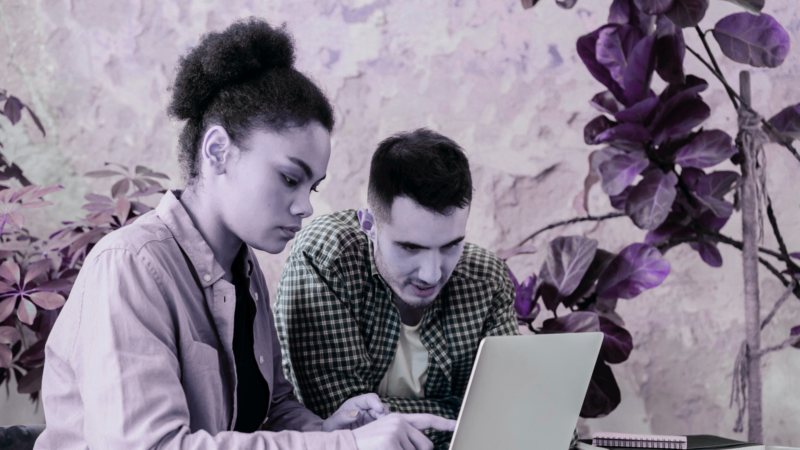 Two people sitting at a desk leaning over a laptop.