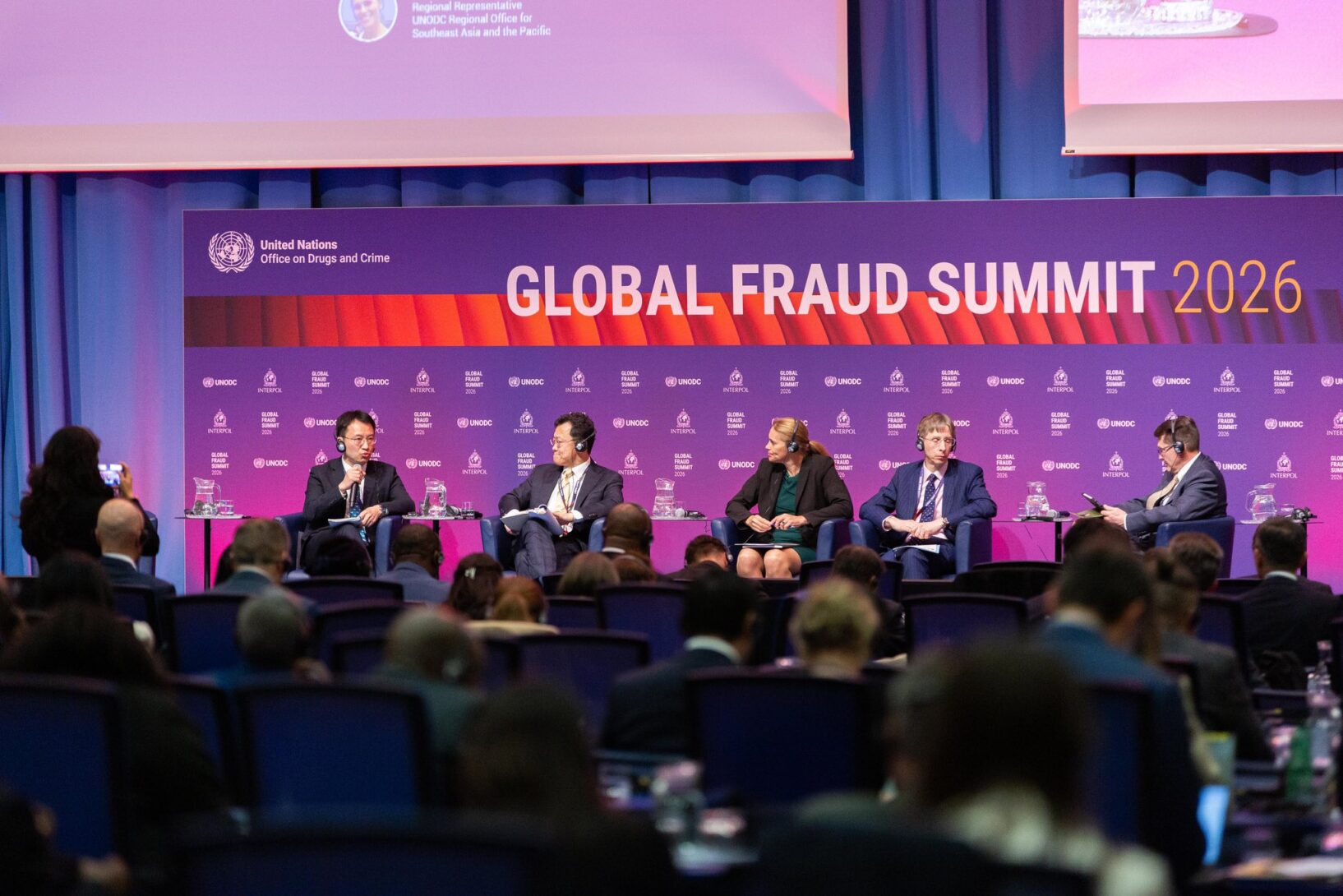 This image shows a panel of speakers speaking before a crowd. A sign in the background says "Global Fraud Summit."