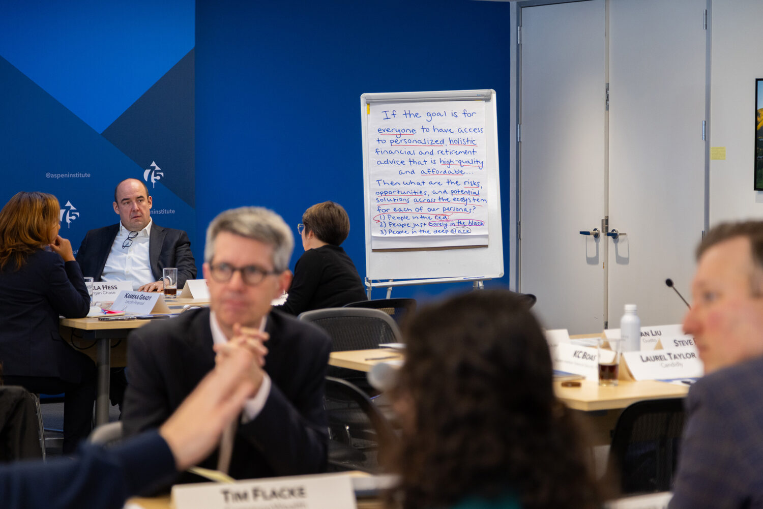 Event attendees sit at tables speaking. In focus behind them, is a flipchart with notes about retirement savings advice.