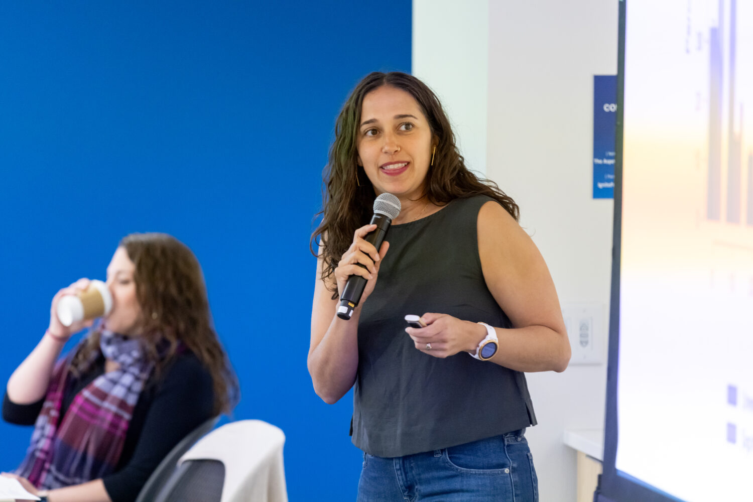 A woman holds a microphone in front of a screen presentation.