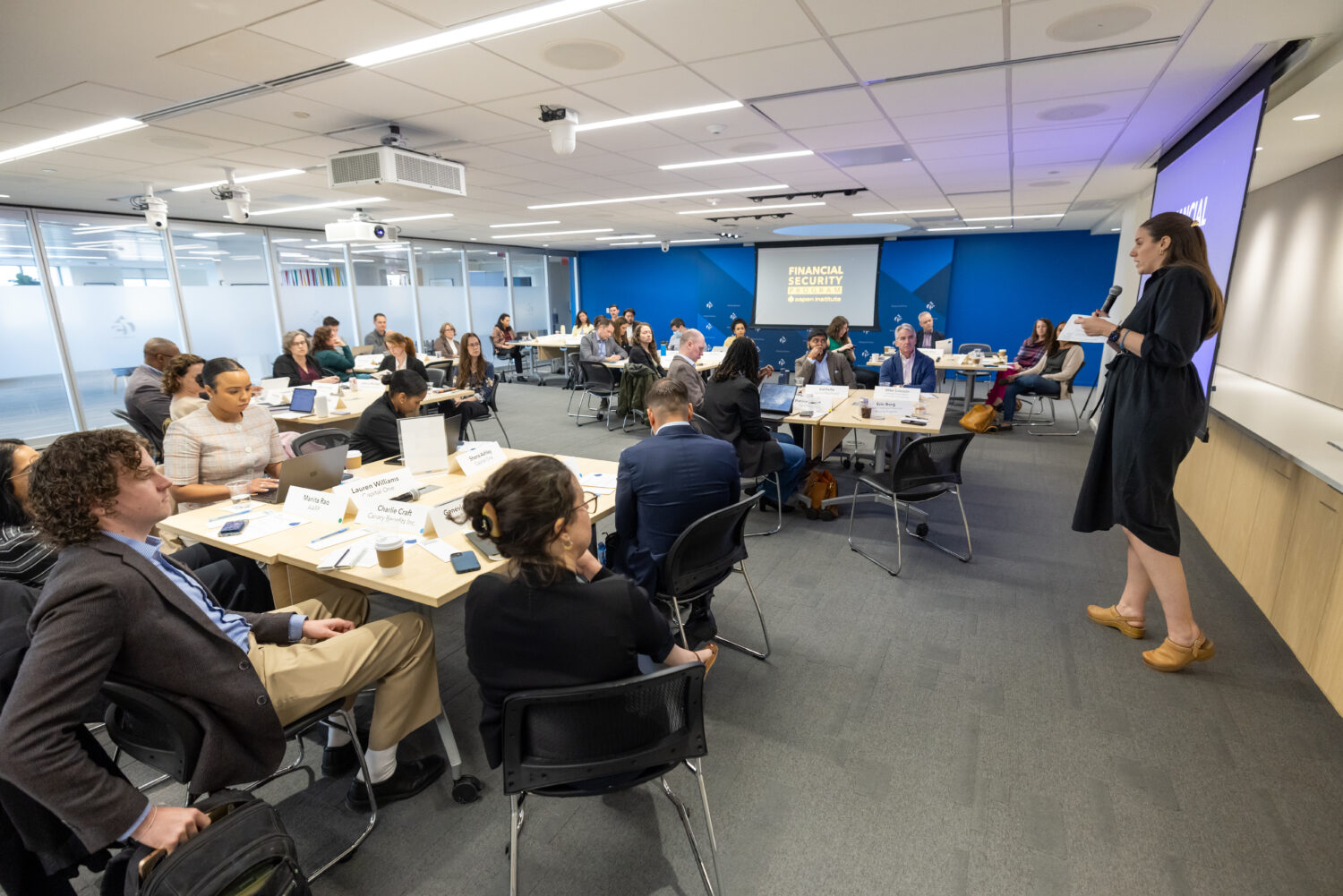 A picture of the full room at an event. People are seated in groups at multiple tables and one person is presenting at the front of the room.