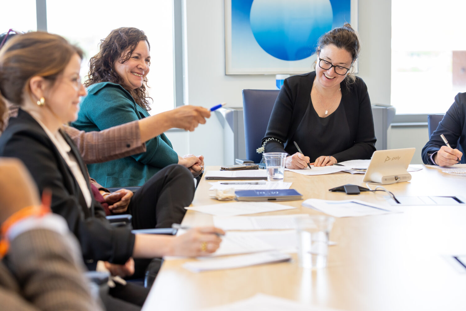 A group of people sit around a table in discussion. One woman is smiling and taking notes.