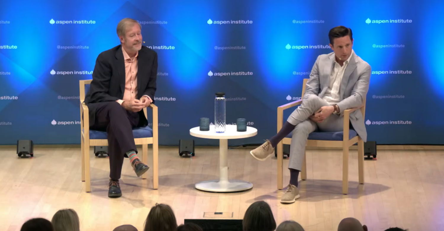 Screen capture of two men seated on a stage in front of an Aspen Institute background. Two glasses and a water carafe sit on a low white table between them. The heads of some audience members can be seen in the foreground. The man on the left is light haired, bearded, and wearing black pants, a salmon-colored dress shirt, and a black jacket. The man on the left is dark-haired, clean shaven, and wearing a grey suit and a white dress shirt.