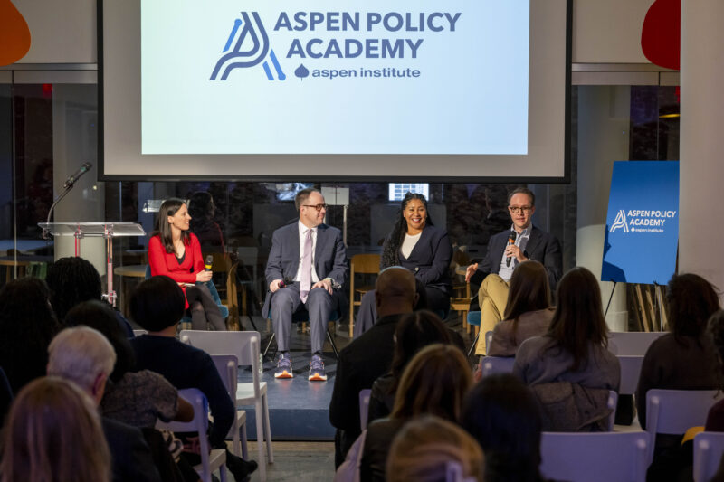 Aspen Policy Academy Director Betsy Cooper and another guest facilitate a panel with former Mayors London Breed and G.T. Bynum. The 4 speakers sit in chairs on stage with the Aspen Policy Academy logo displayed on a projector screen behind them.
